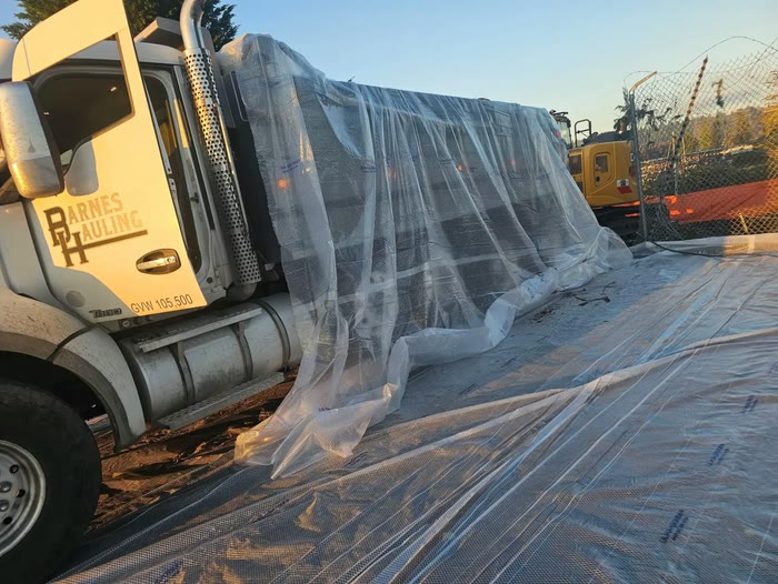 A Barnes Hauling truck with its load wrapped in clear plastic sheeting on a lined site, an excavator behind a fence.