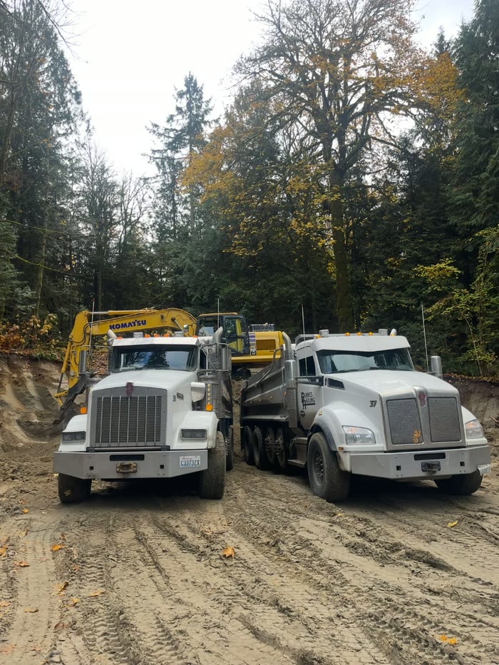 Two Barnes Hauling dump trucks side by side in a freshly dug pit, a yellow Komatsu excavator loading behind them under tall trees.