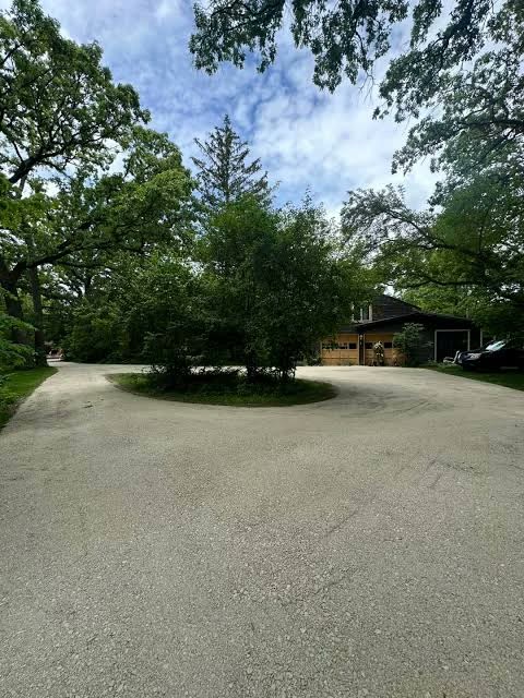 A wide gravel driveway looping around a planted island in front of a house and garage, under tall trees.