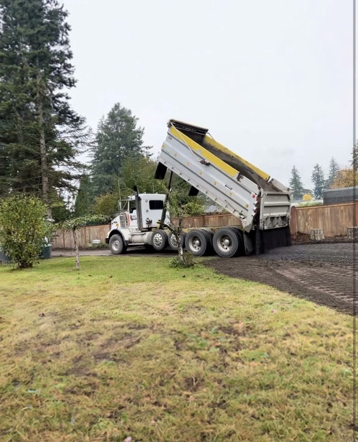 A dump truck with its bed raised, tipping a load along a graded driveway beside a lawn and a wooden fence.