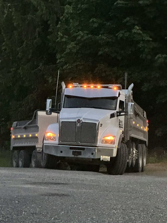 A white Kenworth dump truck with the Barnes Hauling logo on the door, amber cab lights on at dusk.