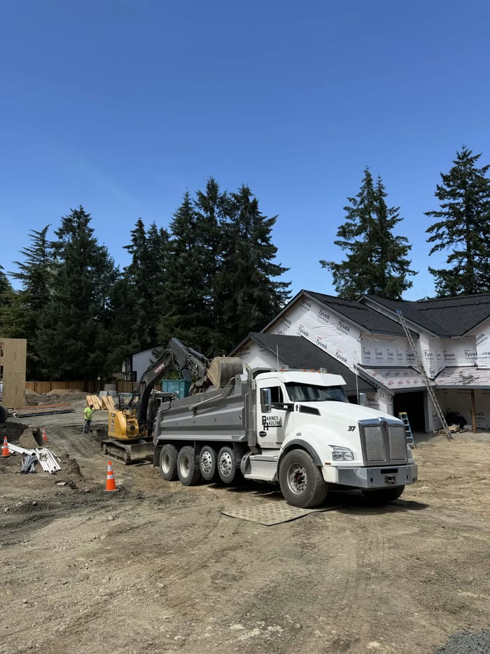 An excavator fills a Barnes Hauling dump truck on a new-home construction site, traffic cones and pipe on the dirt beside it.