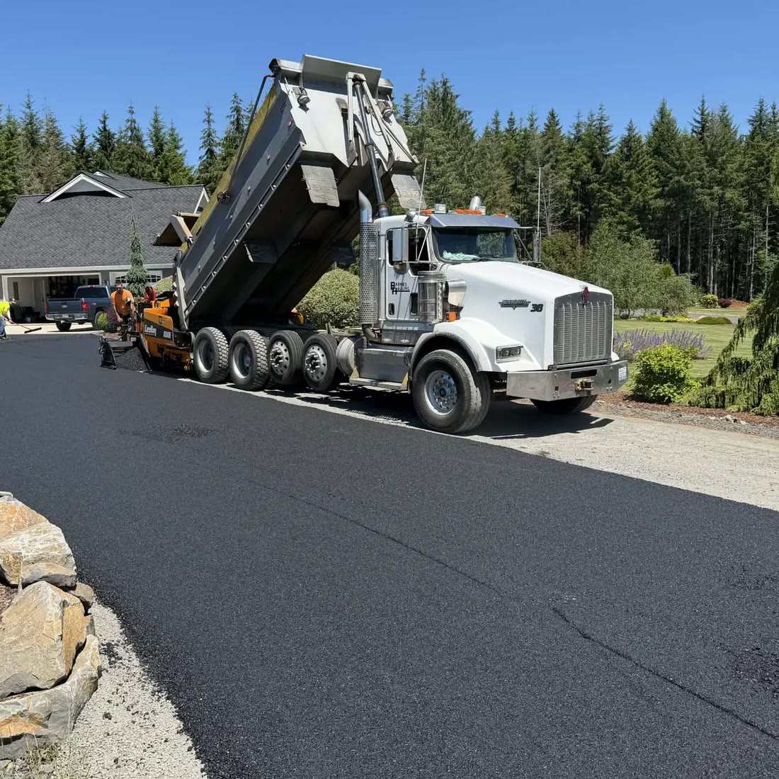 A Barnes Hauling dump truck tips asphalt into a paving machine on a new
                    driveway, with fir trees behind.