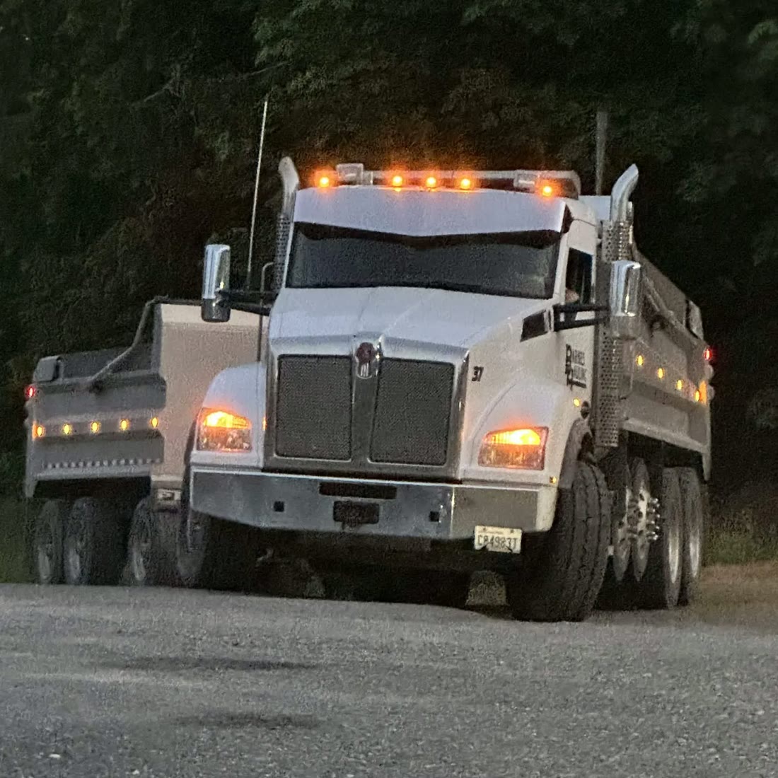 A white Barnes Hauling Kenworth dump truck with its amber cab lights on at dusk,
                    a second truck parked behind it.