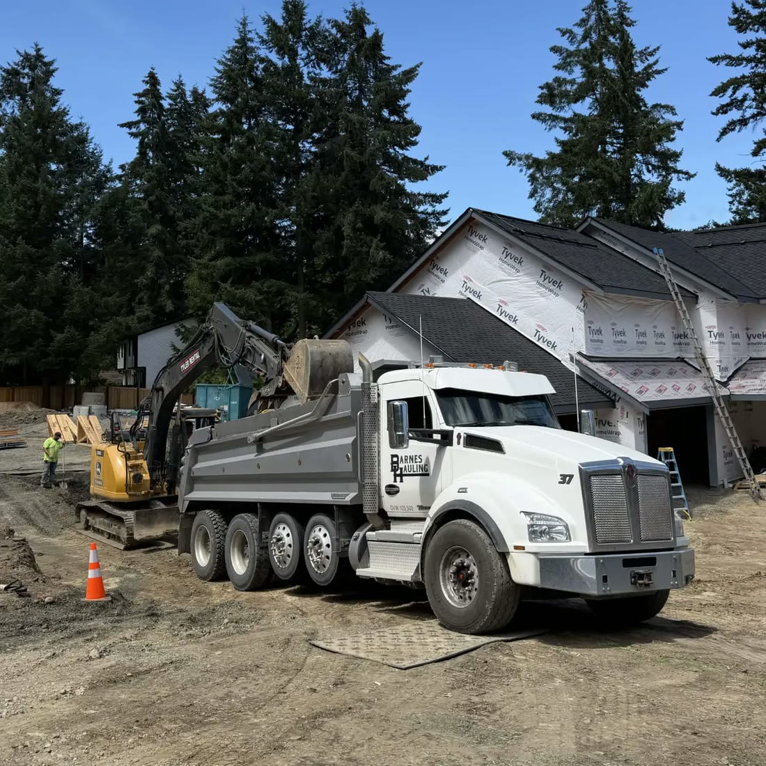 An excavator loads a Barnes Hauling dump truck on a new-home construction
                    site, a worker and traffic cones beside it.
